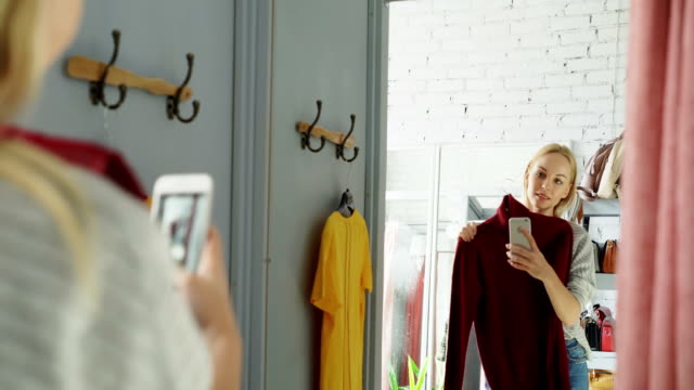 Cliente-femenino-está-haciendo-espejo-selfie-con-nuevo-puente-punto-usando-el-teléfono-inteligente.-Ella-está-de-pie-en-el-montaje-de-sala-en-la-tienda-de-ropa-y-posando-con-sonrisa-encantadora.