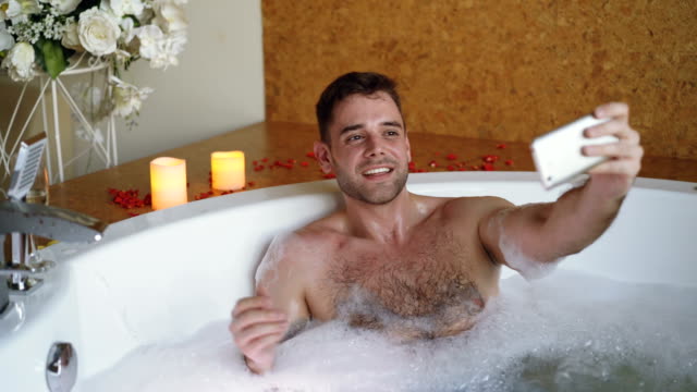 Young-bearded-guy-famous-blogger-is-recording-video-in-hot-tub-in-day-spa-using-smartphone.-Burning-candles,-champagne-glass-and-flowers-are-visible.