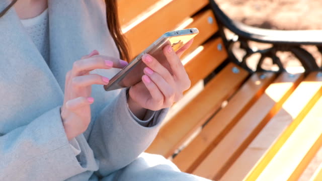 Woman-typing-a-message-in-her-phone-sitting-on-the-bench-in-park.-Close-up-hands.
