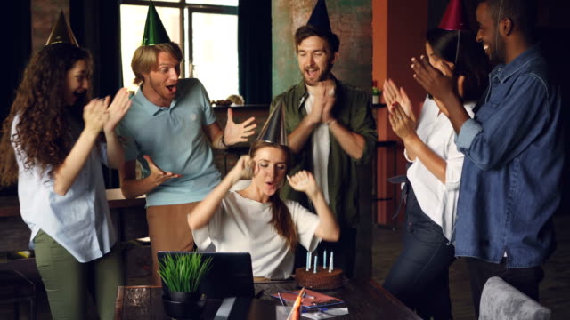 Attractive-young-woman-is-blowing-candles-on-birthday-cake-and-laughing-while-her-coworkers-are-clapping-hands-and-congratulating-her.-Party-and-work-concept.
