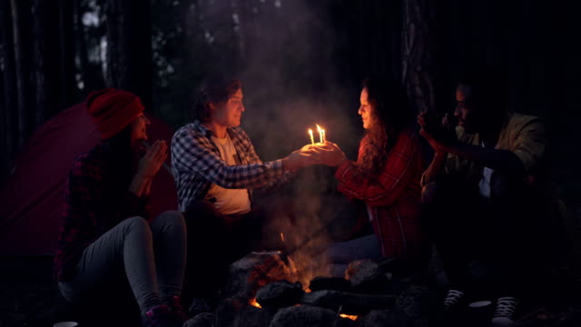 Creative-people-are-congratulating-their-friend-on-birthday-in-forest,-clapping-hands-and-giving-her-cake-while-girl-is-blowing-candles-and-smiling.-Nature-and-celebration-concept.