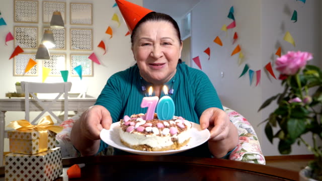 Mujer-senior-feliz-sosteniendo-pastel-a-cámara-celebrando-cumpleaños-con-su-familia-a-través-de-video-chat