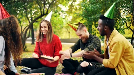 Schönes-Mädchen-in-Partyhut-feiert-Geburtstag-mit-Freunden-im-Park-Picknick-machen-Wunsch,-bläst-Kerzen-auf-Kuchen-und-Spaß-Lachen.-Veranstaltungen-und-Natur-Konzept.
