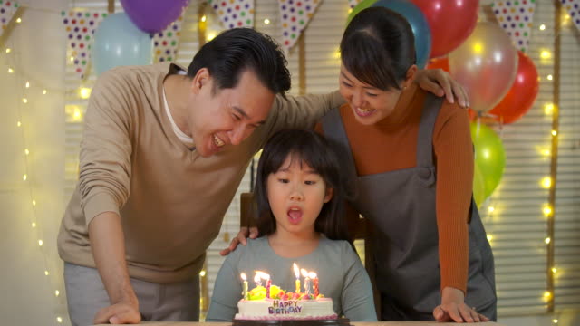 Ein-niedliche-kleine-asiatische-Mädchen-am-Tisch-sitzen-und-bläst-Kerzen-auf-Geburtstagskuchen-während-ihrer-Familie-hinter-und-Sing-ein-Lied-mit-ihr.-Sie-gibt-ein-breites-Lächeln-und-ihre-Familie-begrüßen-sie.-In-Zeitlupe.