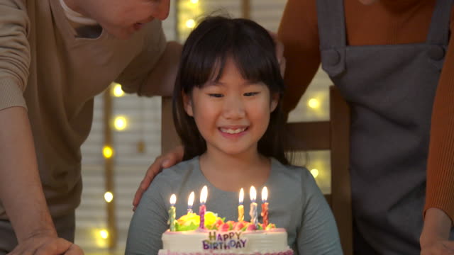 Ein-niedliche-kleine-asiatische-Mädchen-am-Tisch-sitzen-und-bläst-Kerzen-auf-Geburtstagskuchen-während-ihrer-Familie-hinter-und-Sing-ein-Lied-mit-ihr.-Sie-gibt-ein-breites-Lächeln-und-ihre-Familie-begrüßen-sie.-In-Zeitlupe.