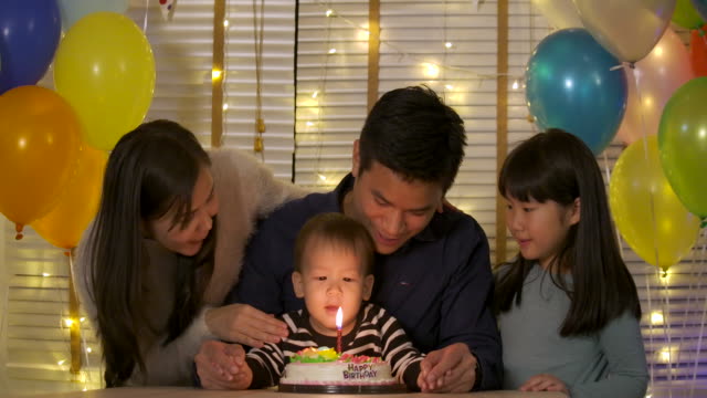 A-cute-little-boy-sitting-at-table-and-blowing-candles-on-birthday-cake-while-his-family-standing-behind-and-sing-a-song-to-him.-In-slow-motion.