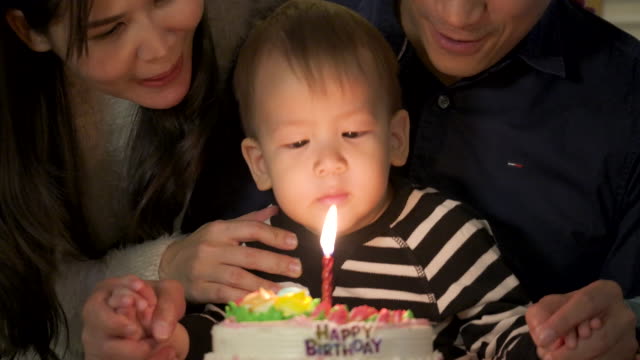 A-cute-little-boy-sitting-at-table-and-blowing-candles-on-birthday-cake-while-his-family-standing-behind-and-sing-a-song-to-him.-In-slow-motion.