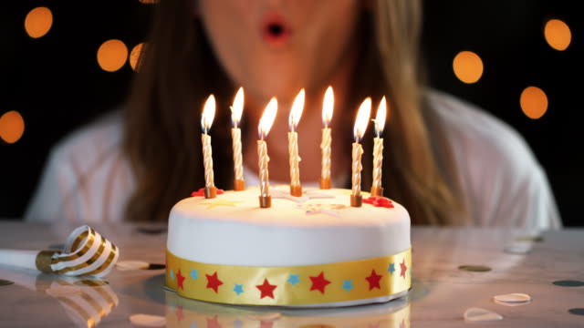 Close-up-of-a-woman-blowing-out-seven-lit-candles-on-a-white,-decorated-birthday-cake,-a-party-blower-beside-it,-bokeh-lights-in-the-background