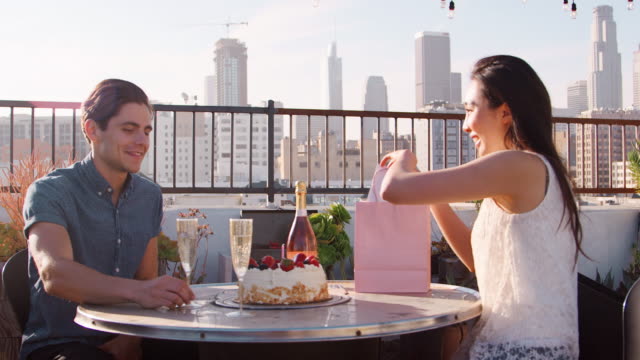 Mann-Frau-Geschenk-und-Karte-zu-geben,-wie-sie-auf-Dachterrasse-mit-Skyline-der-Stadt-im-Hintergrund-feiern