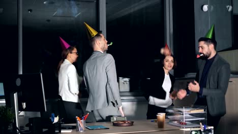 Happy-young-people-office-workers-are-dancing-at-company-party-wearing-party-hats-and-blowing-horns.-Birthday-cake-and-gift-box-are-visible-on-desk.