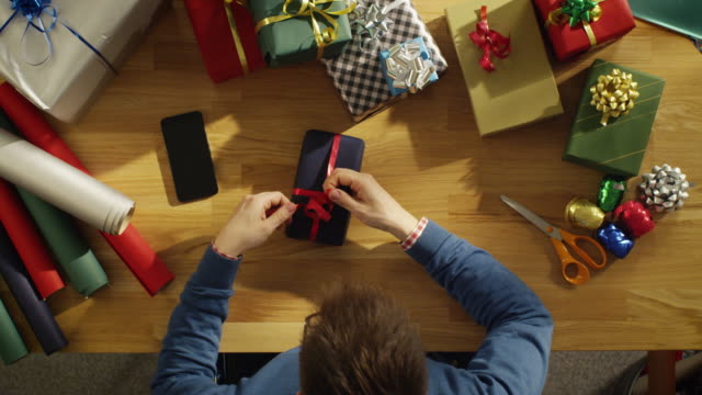 Draufsicht-der-junge-Mann-gekonnt-Verpackung-bunte-Geschenke-im-sonnigen-Atelier.-Smartphone-mit-grünen-Mock-up-Bildschirm-liegt-auf-dem-Tisch.