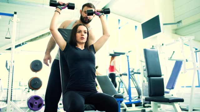 Trainer-helps-young-strong-brunette-woman-doing-exercise-in-fitness-club-and-gym-center