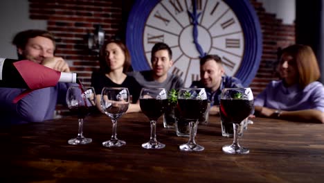 Close-up-shot-of-the-sommelier-who-pours-a-red-wine-on-a-glass-for-a-company-of-friends-who-sits-in-the-background-and-enjoy-a-neat-process