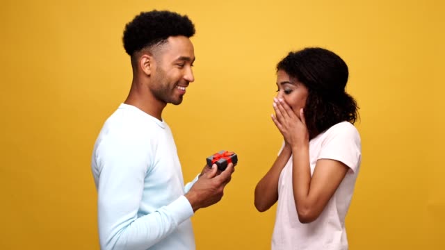 Smiling-young-african-man-giving-a-present-box-to-his-girlfriend-isolated-over-yellow-background