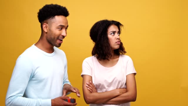 Smiling-young-african-man-giving-a-present-box-to-his-upset-grumpy-girlfriend-isolated-over-yellow-background