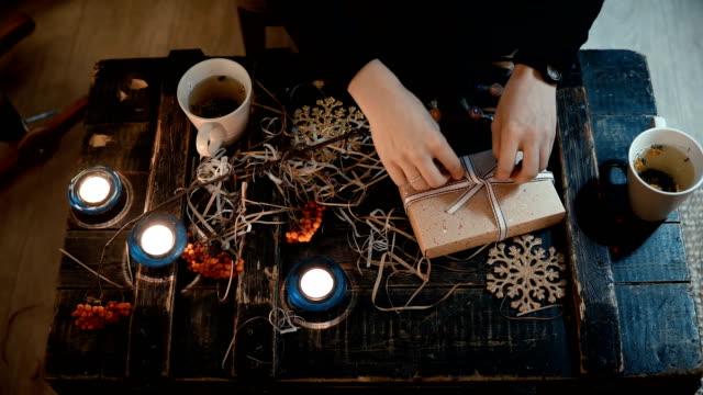 Top-close-up-view-of-female-hands-making-gift-sitting-at-the-table-at-home.-Woman-binds-a-Christmas-box-with-a-ribbon