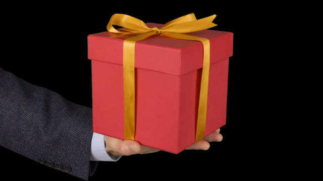 Mans-hand-in-a-business-suit-holds-a-red-gift-box-with-a-gold-bow.-Hand-with-a-gift-appears-in-the-frame.-Red-gift-box.-A-businessman-gives-a-gift.-Alpha-channel-chroma-key-transparent-background.
