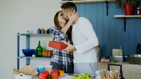 Laughing-attractive-woman-surprising-his-boylfriend-with-a-gift-at-home-in-the-kitchen-while-he-cooking-breakfast