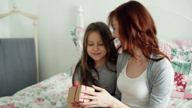 Glücklich-Younf-Mutter-küssen-ihre-süße-Tochter-präsentiert-Geschenkbox-auf-Feier-auf-in-hellen-Schlafzimmer-zu-Hause-sitzen