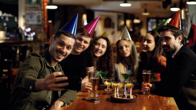 Happy-young-people-in-birthday-party-hats-are-recording-video-with-smartphone-sitting-at-table-in-bar.-They-are-posing,-laughing-and-clapping-hands.