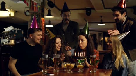 Young-pretty-brunette-is-making-wish-and-blowing-out-candles-on-birthday-cake-while-celebrating-birthday-in-cafe-with-friends.-Happy-people-in-party-hats-are-clapping-hands.