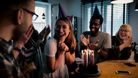 Happy-Caucasian-girl-makes-a-wish-at-birthday-cake-with-candles.-Multiethnic-friends-at-surprise-party-slow-motion