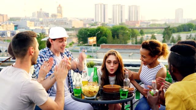 Young-woman-birthday-girl-is-making-wish,-blowing-candle-on-cake-and-clapping-hands-while-her-friends-are-congratulating-her-and-clinking-bottles-with-drinks-during-rooftop-party.
