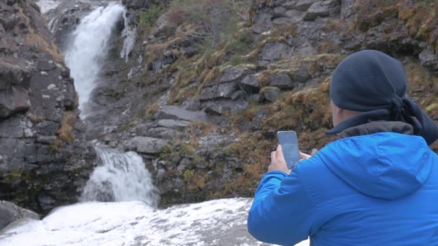Ein-Mann-fotografiert-einen-schönen-Wasserfall-in-den-Bergen,-auf-einem-Smartphone.-Führt-ein-aktives-und-interessantes-Leben-und-stellt-Fotos-von-ihren-Reisen-in-den-sozialen-Netzwerken.