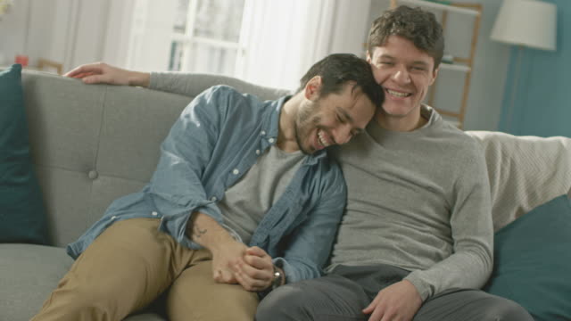 Cute-Attractive-Male-Gay-Couple-Sit-Together-on-a-Sofa-at-Home.-Boyfriends-are-Hugging-and-Embracing-Each-Other.-They-are-Joyful-and-Laughing.-They-are-Casually-Dressed-and-Room-Has-Modern-Interior.