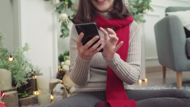 Cheerful-happy-young-Asian-woman-using-smartphone-to-check-social-media-in-her-living-room-at-home-in-Christmas-Festival.-Lifestyle-women-celebrate-Christmas-and-New-year-concept.