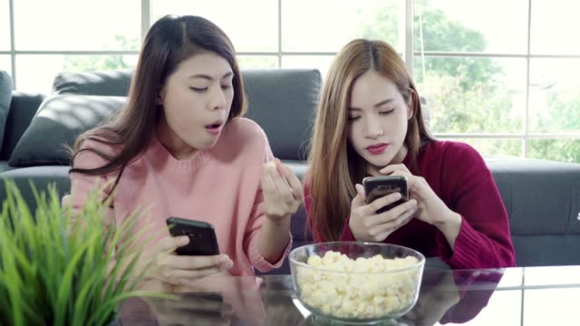 Asian-women-using-smartphone-and-eating-popcorn-in-living-room-at-home,-group-of-roommate-friend-enjoy-funny-moment-while-lying-on-the-sofa.-Lifestyle-women-relax-at-home-concept.