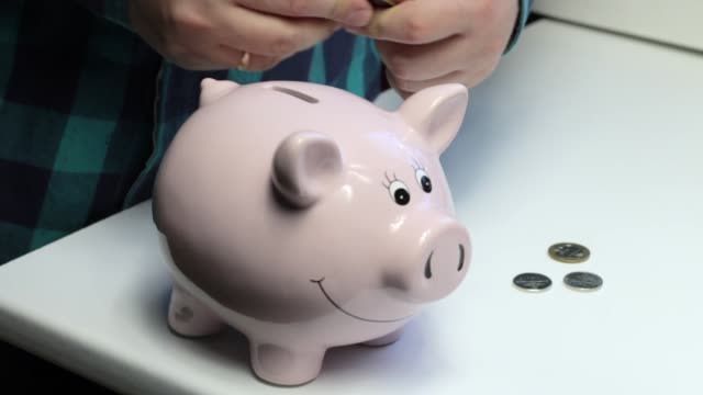 On-the-table-there-is-a-piggy-bank-in-the-form-of-a-pink-pig.-Lying-scattered-coins.-A-man-collects-coins-from-the-table-and-puts-them-in-a-piggy-bank.