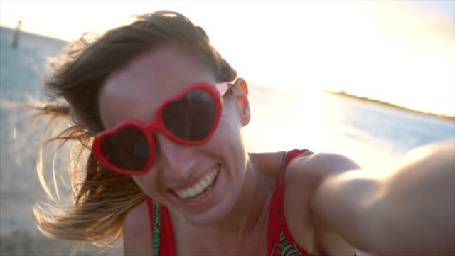 Mujer-joven-con-el-corazón-en-forma-de-gafas-de-sol-rojo-tomar-selfie-fotografía-al-atardecer-en-la-hermosa-playa-en-Hawai.-Cámara-lenta