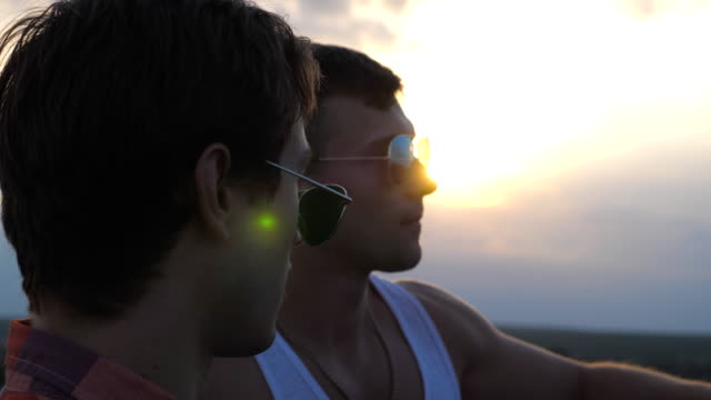 Profile-of-young-male-couple-in-sunglasses-standing-on-the-edge-of-rooftop-and-talking.-Handsome-gay-boys-relaxing-on-roof-of-high-rise-building-and-enjoying-beautiful-cityscape.-Close-up-Slow-motion