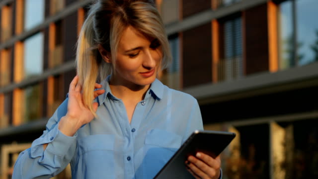 Close-up-portrait-of-a-beautiful-business-woman-holding-and-using-tablet-outside,-girl-looking-at-camera.-Modern-architecture-building-background