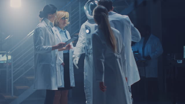 Diverse-Team-of-Aerospace-Scientists-and-Engineers-Wearing-White-Coats-have-Discussion,-Use-Computers,-Design-New-Space-Suit-Adapted-for-Galaxy-Exploration-and-Travel.-Constructing-Astronaut-Suit