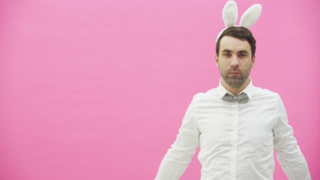 Young-man-standing-on-a-pink-background.-During-this-he-looks-at-the-camera-and-adjusts-the-butterfly-in-his-shirt.-Lays-a-hand-on-the-hand.-The-guy-wears-a-bunny-ear.