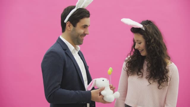 Young-sexy-couple-on-pink-background.-With-hackneyed-ears-on-the-head.-During-this-man-gives-a-soft-toy-hare-and-colored-decorative-eggs-to-his-wife.-Having-kissed-looking-at-the-camera.