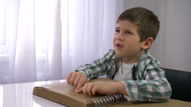Blinde-Kinderjunge,-die-Braille-Buch-mit-Symbolschrift-für-Sehbehinderte-am-Tisch-liest