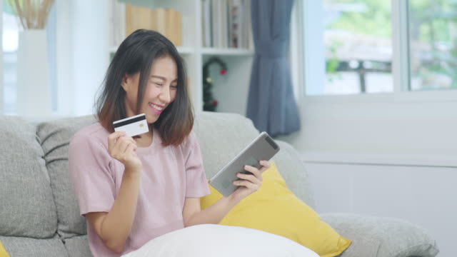 Young-smiling-Asian-woman-using-tablet-buying-online-shopping-by-credit-card-while-lying-on-sofa-when-relax-in-living-room-at-home.-Lifestyle-latin-and-hispanic-ethnicity-women-at-house-concept.