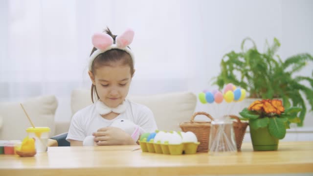 Nice,-cute-girl-is-having-fun-paiting-with-Easter-bunny,-Adorable-girl-is-lulling-bunny-and-cudding-it.-Girl-with-a-beauty-spot-at-her-face-and-is-smiling-gently,-sitting-at-the-wooden-table-with-Easter-decorations.