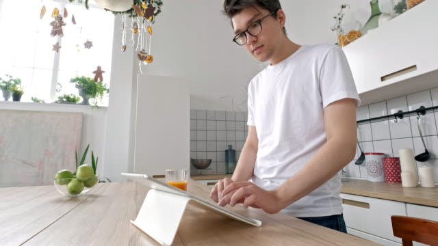 Hombre-atractivo-en-casa-usando-la-tableta-en-la-cocina-enviando-mensaje-en-las-redes-sociales-sonriendo-disfrutando-del-estilo-de-vida-moderno-que-llevaba-camisa-blanca