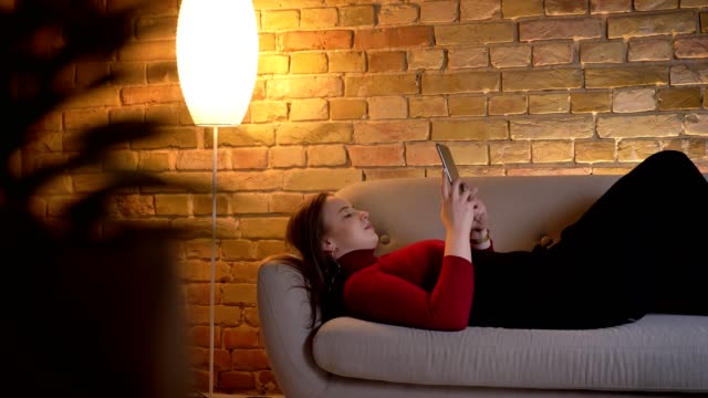 Nahaufnahme-von-jungen-ziemlich-kaukasischen-weiblichen-TEXTen-auf-dem-Tablet,-während-auf-der-Couch-in-innenräumen-liegen