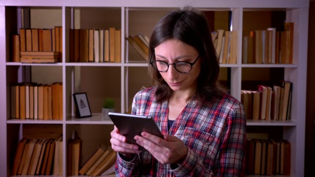 Closeup-shoot-of-young-attractive-female-student-in-glasses-using-the-tablet-and-looking-at-camera-smiling-in-the-university-library-indoors
