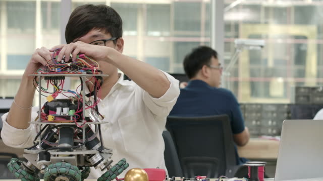 Junge-asiatische-Elektronik-Entwicklungsingenieure-arbeiten-mit-Robotern-und-messen-das-Signal-in-den-elektrischen-Schaltungen-des-Roboterprototyps-in-der-Werkstatt.-Menschen-mit-Technologie--oder-Innovationskonzept.