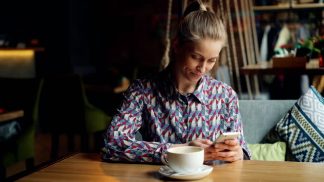 Nettes-Mädchen-mit-Smartphone-in-Café-berühren-Bildschirm-lächelnd-sitzend-am-Tisch