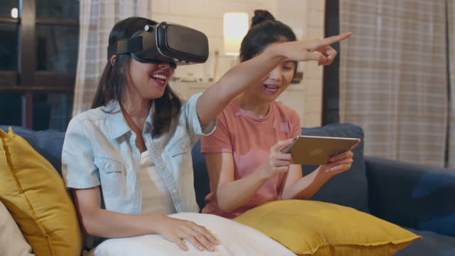 Lesbian-lgbt-women-couple-using-tablet-at-home,-Asian-female-feeling-happy-using-laptop-and-VR-playing-games-together-while-lying-sofa-in-living-room-in-night.-Lover-celebrate-holiday-concept.