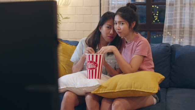 Lesbian-lgbt-women-couple-watching-movie-at-home,-Asian-female-lover-eating-popcorn-scary-and-shock-while-looking-horror-film-together-on-sofa-in-living-room-in-night-concept.-Slow-motion-shot.