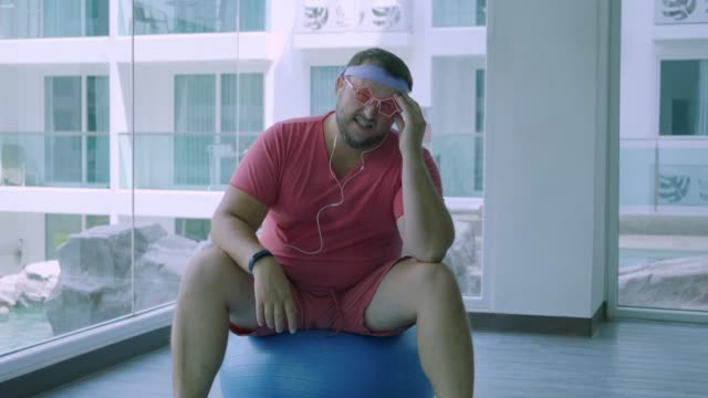 Funny-fat-male-in-pink-glasses-and-in-a-pink-t-shirt-is-engaged-on-a-fit-ball-in-the-gym-depicting-a-girl.-A-freak-man-has-a-headache-on-the-ball-in-the-gym