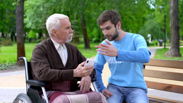 Young-person-explaining-disabled-pensioner-how-to-use-smartphone-outdoors